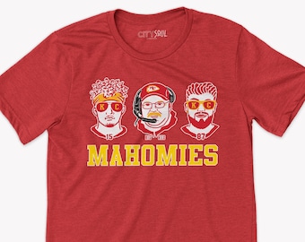 Può includere: Maglietta rossa con una grafica di tre volti di uomini che indossano occhiali da sole e il testo "MAHOMIES". Gli uomini indossano abiti rossi, bianchi e gialli. Il testo è in giallo.