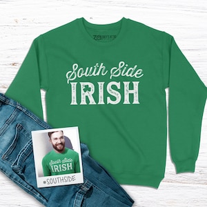 May include: A green sweatshirt with the text "South Side Irish" in white with a shamrock graphic. The sweatshirt is on a white wooden surface with a pair of blue jeans and a photo of a person wearing the same sweatshirt.