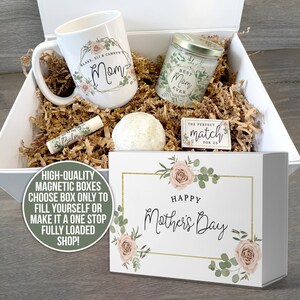 K&ouml;nnte beinhalten: Eine wei&szlig;e Geschenkbox mit magnetischem Verschluss. Die Box ist gef&uuml;llt mit einer wei&szlig;en Tasse mit Blumenmuster und dem Text "Mama", einer Kerze mit dem Text "Beste Mama der Welt", einer Badebombe, einem Lippenbalsam und einem kleinen Holzschild mit dem Text "Die perfekte &Uuml;bereinstimmung f&uuml;r uns". Die Box ist mit einem Blumenmuster und dem Text "Frohen Muttertag" verziert.