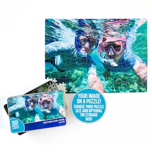 May include: A 300-piece jigsaw puzzle in a metal tin with a photo of a couple scuba diving. The puzzle is titled "Honeymoon Scuba Diving June 2021". The tin has a blue background with white text and a white border. The text on the tin reads "Your image on a puzzle! Choose your puzzle size and optional tin storage box!"