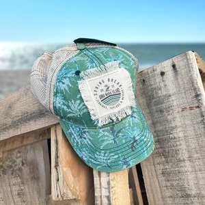May include: A green and blue trucker hat with a white mesh back. The hat has a palm tree print and a patch that says "Spring Break 2022 Anna Maria Island Jennings, Taylors & Peters".