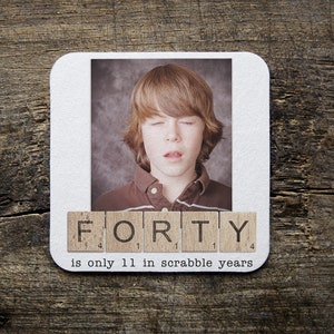 May include: A white coaster with a photo of a young person with brown hair and a brown shirt. The photo is surrounded by Scrabble tiles that spell out "FORTY". The text below the tiles reads "is only 11 in Scrabble years".