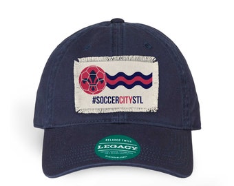 Op de afbeelding: Een donkerblauwe baseballpet met een verweerde stoffen patch met een voetbal en de tekst "#SOCCERCITYSTL". De pet heeft een groen label met de tekst "RELAXED TWILL LEGACY".