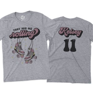 Puede incluir: Camiseta gris con el texto "They See Me Rolling" y un par de patines rosas con ruedas arco&iacute;ris. La parte posterior de la camiseta tiene el texto "Kelsey 11" en una fuente retro.