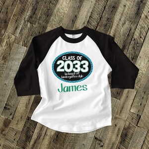 May include: A white and black baseball tee shirt with a blue and teal circle graphic that says "Class of 2033 kicking it off kindergarten style" and the name "James" in green text.