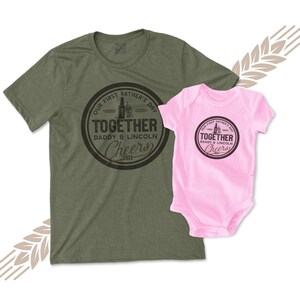 May include: A green t-shirt with a black circle graphic that says "Our First Father's Day Together Daddy & Lincoln Cheers 2023". A pink baby onesie with the same graphic is shown next to the t-shirt.