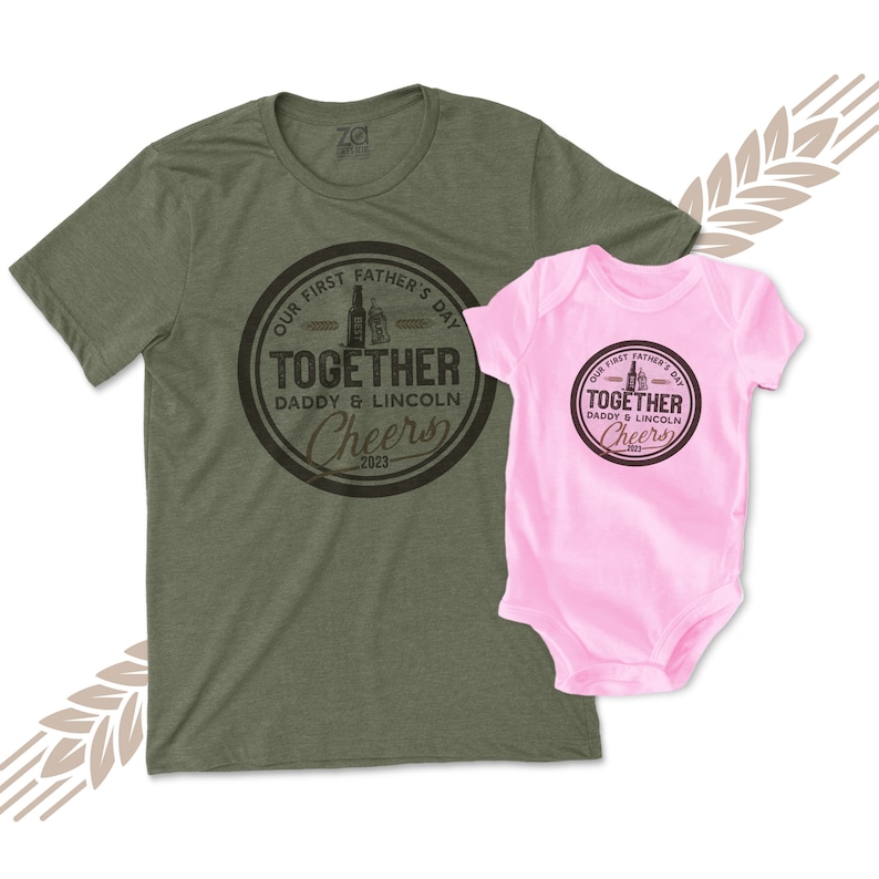 May include: A green t-shirt with a black circle graphic that says "Our First Father's Day Together Daddy & Lincoln Cheers 2023". A pink baby onesie with the same graphic is shown next to the t-shirt.