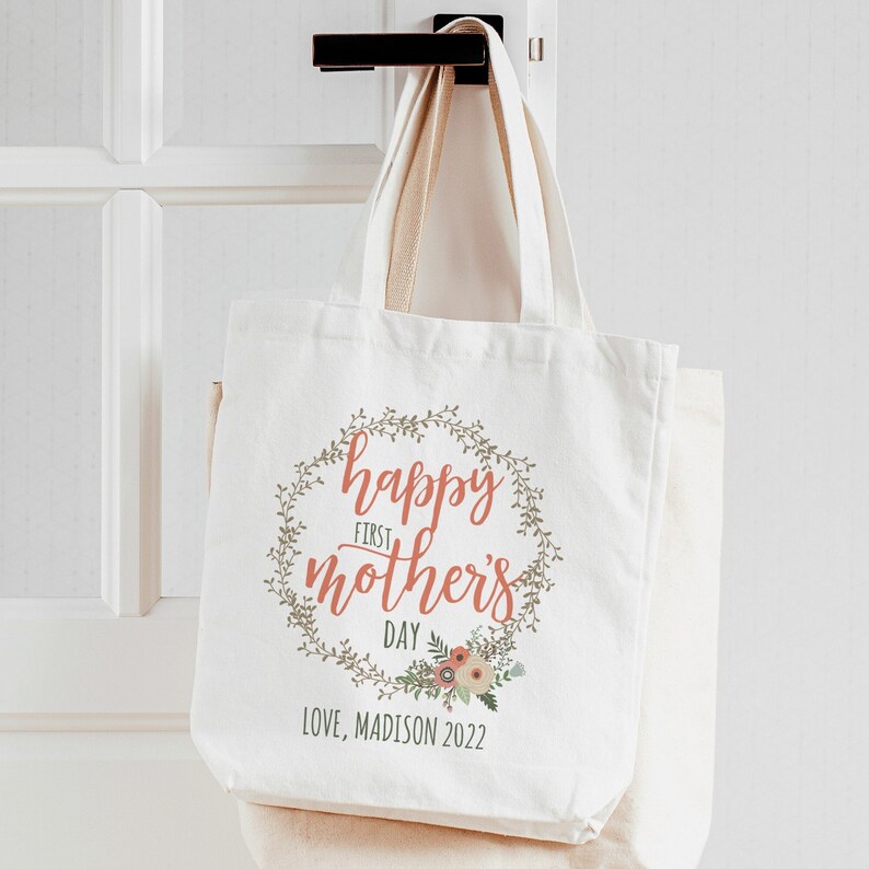 May include: White canvas tote bag with a floral wreath design. The wreath has green leaves and pink flowers. The text "happy first mother's day" is in a pink script font. The text "LOVE, MADISON 2022" is below the wreath.