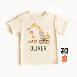 May include: A beige short-sleeved t-shirt with a watercolor illustration of a yellow excavator digging up pumpkins. The text "Oliver" is printed below the excavator. The shirt has a small logo in the bottom right corner that says "ZOEY'S ATTIC" and "TOP SELLER Etsy SINCE 2009".
