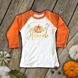 Pode incluir: Camiseta branca de manga comprida com mangas laranja. A camiseta tem um design de ab&oacute;bora e folhas com o nome "Hannah" em letras douradas.