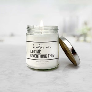 May include: A white candle in a clear glass jar with a silver lid. The candle has a black label with the text "hold on LET ME OVERTHINK THIS" and the text "HAND POURED IN ST. LOUIS, MO 8 OZ | 100% SOY WAX".