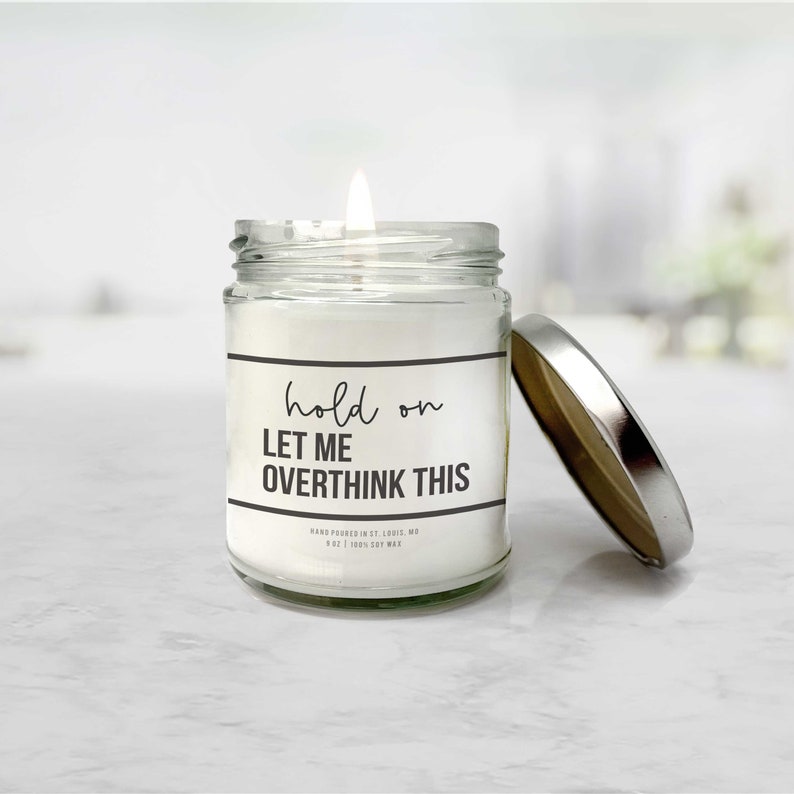May include: A white candle in a clear glass jar with a silver lid. The candle has a black label with the text "hold on LET ME OVERTHINK THIS" and the text "HAND POURED IN ST. LOUIS, MO 8 OZ | 100% SOY WAX".