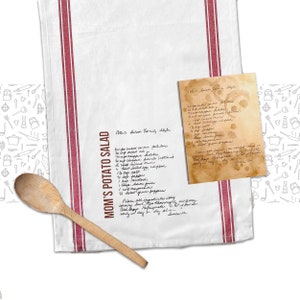 May include: A white tea towel with red stripes and a handwritten recipe for "Mom's Potato Salad" and "Polaris Salad Family Style". The recipe includes ingredients like celery, potatoes, and mayonnaise. The towel is decorated with a pattern of kitchen utensils.