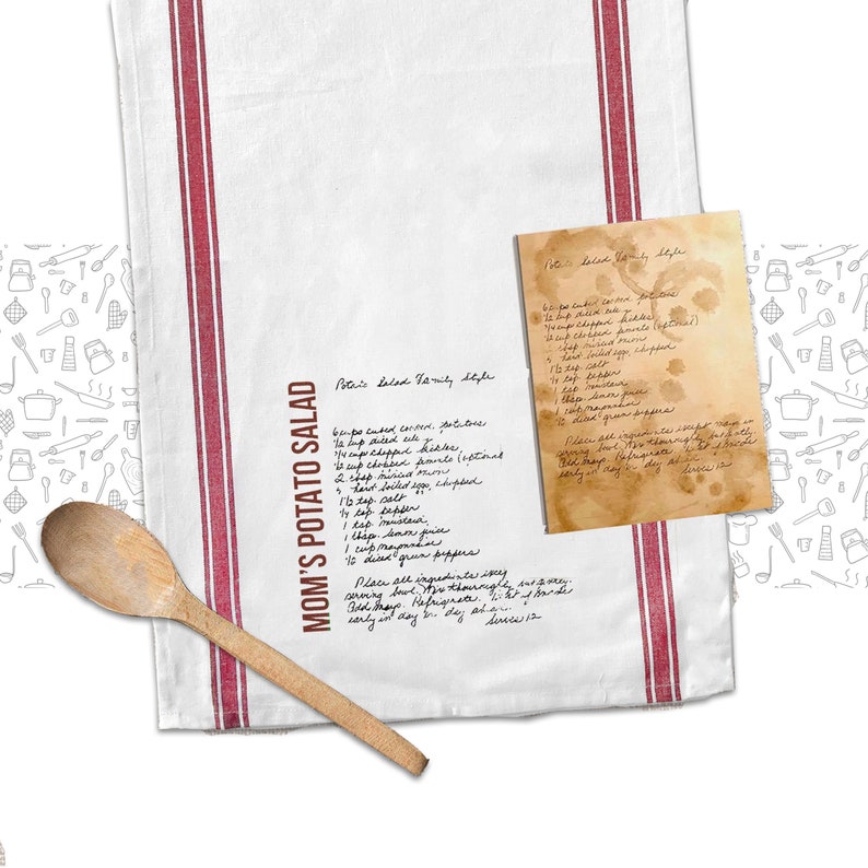 May include: A white tea towel with red stripes and a handwritten recipe for "Mom's Potato Salad" and "Polaris Salad Family Style". The recipe includes ingredients like celery, potatoes, and mayonnaise. The towel is decorated with a pattern of kitchen utensils.