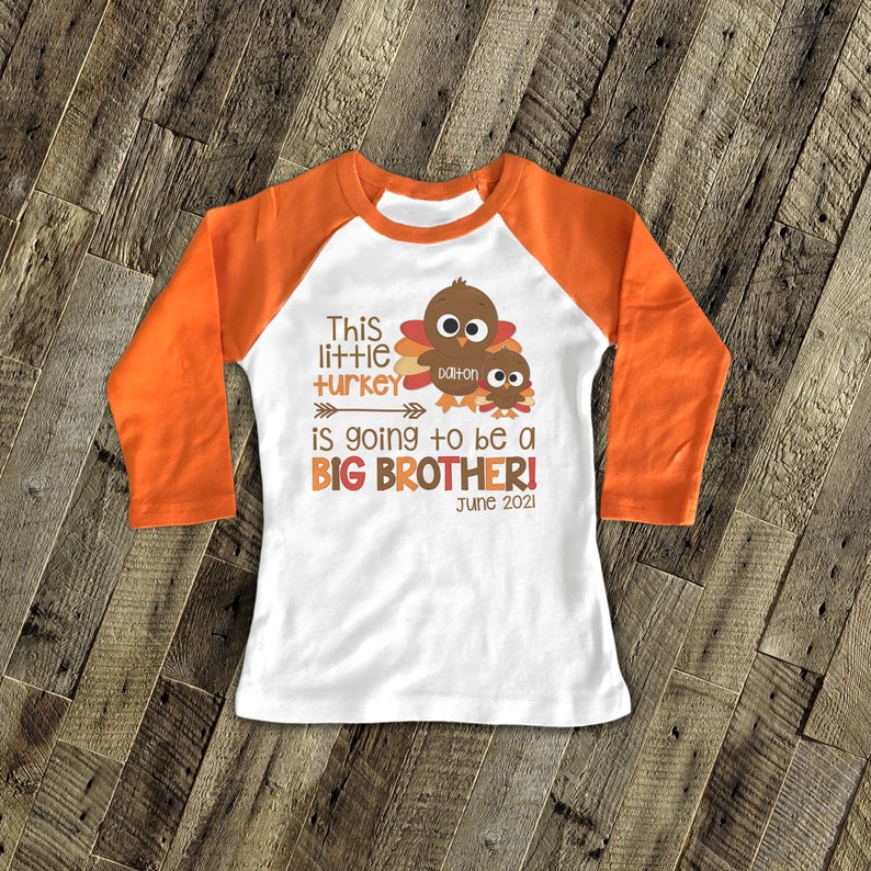 K&ouml;nnte beinhalten: Wei&szlig;es Langarmshirt mit orangenen &Auml;rmeln. Das Shirt hat ein Truthahn-Motiv mit dem Text "This little turkey is going to be a BIG BROTHER! June 2021" und dem Namen "Dalton".