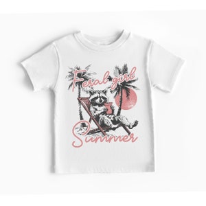 May include: White t-shirt with a graphic of a raccoon sitting in a beach chair with a drink, palm trees, and the text "Feral girl summer" in pink.