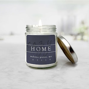 May include: A glass jar candle with a white label that says "cozy HOME" and "webster groves, mo 63119". The label is a dark blue color with white text. The candle is lit and the lid is off.