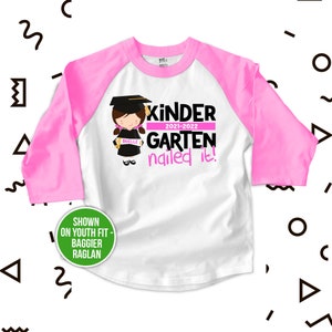 Op de afbeelding: Roze en wit baseballshirt voor kinderen met een roze en zwarte afbeelding van een meisje dat een afstudeerpet en -jurk draagt en een diploma vasthoudt. De tekst op het shirt luidt "Kinder 2021-2022 Garten nailed it!"