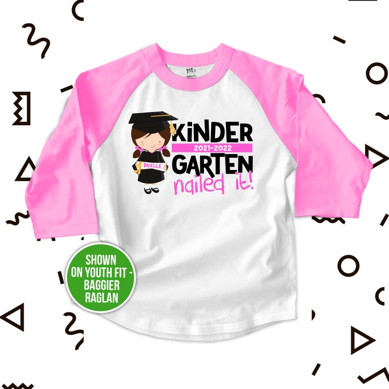 Op de afbeelding: Roze en wit baseballshirt voor kinderen met een roze en zwarte afbeelding van een meisje dat een afstudeerpet en -jurk draagt en een diploma vasthoudt. De tekst op het shirt luidt "Kinder 2021-2022 Garten nailed it!"