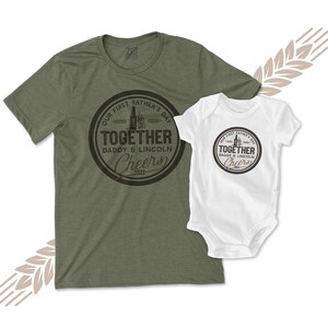 May include: A green t-shirt and a white onesie with a brown circle design that says "Our First Father's Day Together Daddy & Lincoln Cheers 2023".