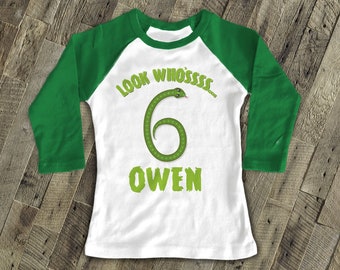 May include: A white and green long-sleeved baseball t-shirt with a green snake design. The shirt says "Look Who's... 6 Owen".