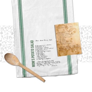May include: A white kitchen towel with green stripes and a handwritten recipe for "Mom's Potato Salad" on it. The recipe includes ingredients like potatoes, celery, onions, mayonnaise, and mustard. The towel also has a handwritten recipe for "Polaris Salad Family Style" on a separate piece of paper.