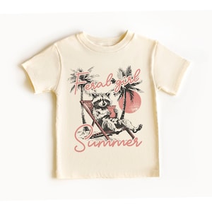 May include: A cream colored t-shirt with a graphic of a raccoon sitting in a lounge chair with a drink. The text "Feral girl summer" is printed in pink on the shirt.