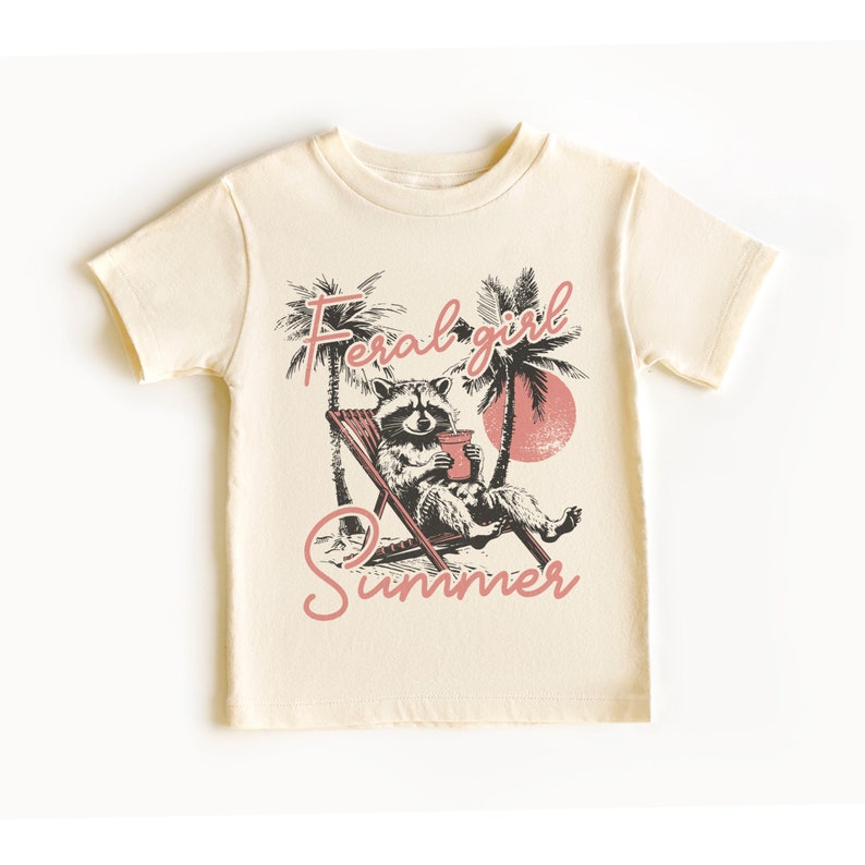 May include: A cream colored t-shirt with a graphic of a raccoon sitting in a lounge chair with a drink. The text "Feral girl summer" is printed in pink on the shirt.