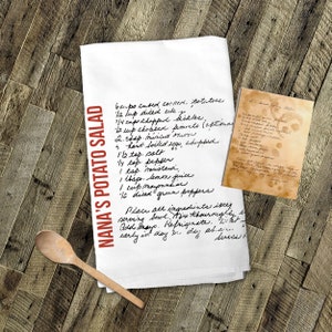 May include: A white kitchen towel with a handwritten recipe for Nana's Potato Salad. The recipe includes ingredients like potatoes, celery, pickles, mayonnaise, and green peppers. The towel is folded in half and has a wooden spoon next to it.