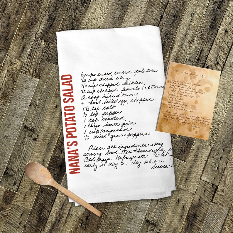 May include: A white kitchen towel with a handwritten recipe for Nana's Potato Salad. The recipe includes ingredients like potatoes, celery, pickles, mayonnaise, and green peppers. The towel is folded in half and has a wooden spoon next to it.