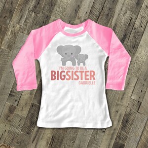 May include: A white and pink long-sleeved baseball tee shirt with a grey elephant and a smaller grey elephant holding hands. The shirt says "I'm going to be a big sister Gabrielle".