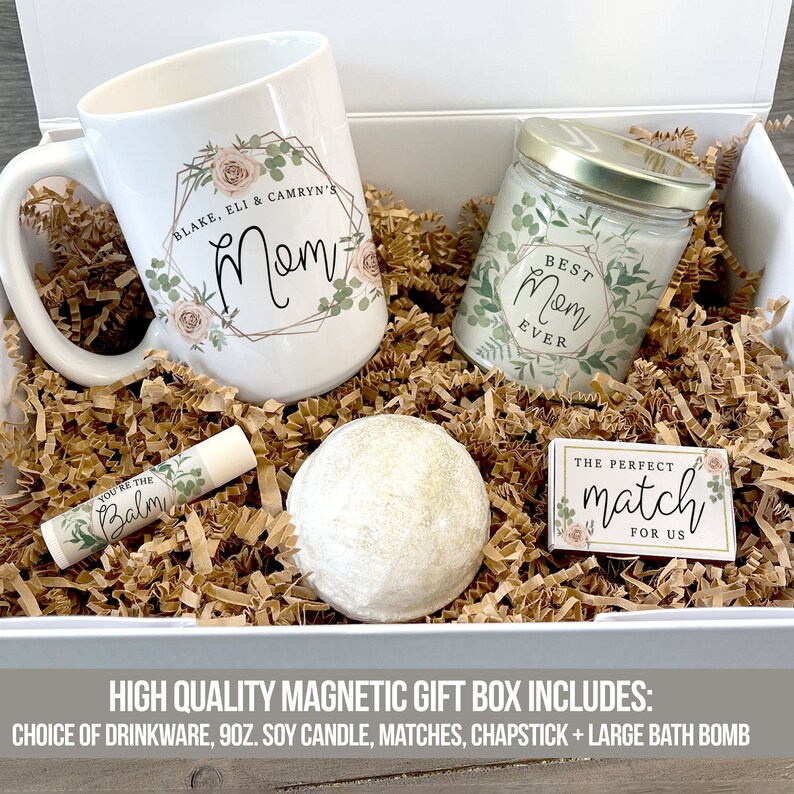 K&ouml;nnte beinhalten: Eine wei&szlig;e Geschenkbox gef&uuml;llt mit braunem Seidenpapier. Die Box enth&auml;lt eine wei&szlig;e Tasse mit einem Blumenmuster und dem Text "Blake, Eli & Camryn's Mom", ein klares Glasgef&auml;&szlig; mit einer Kerze und einem Blumenmuster und dem Text "Best Mom Ever", eine wei&szlig;e Badebombe, eine kleine Tube Lippenbalsam mit dem Text "You're the Balm" und eine kleine wei&szlig;e Box mit einem Blumenmuster und dem Text "The Perfect Match for Us".