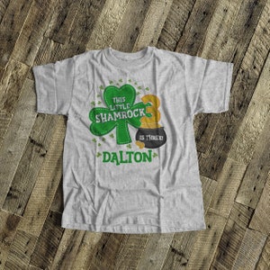 K&ouml;nnte beinhalten: Ein graues T-Shirt mit einem gr&uuml;nen Kleeblatt und dem Text "This Little Shamrock is Three!" Das Shirt hat auch einen Topf mit Gold mit der Nummer "3" darauf und den Namen "DALTON" untenaufgedruckt.