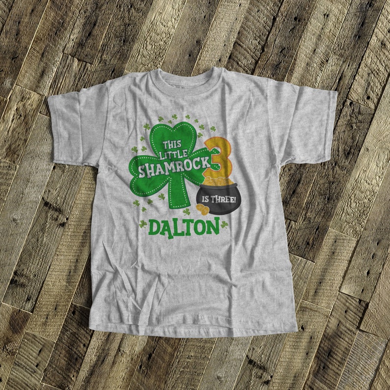 K&ouml;nnte beinhalten: Ein graues T-Shirt mit einem gr&uuml;nen Kleeblatt und dem Text "This Little Shamrock is Three!" Das Shirt hat auch einen Topf mit Gold mit der Nummer "3" darauf und den Namen "DALTON" untenaufgedruckt.