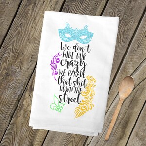 May include: White kitchen towel with a Mardi Gras theme. It features a purple, green, and gold design with a mask and the text "We don't hide our crazy we parade that shit down the street."