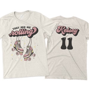 Puede incluir: Dos camisetas de color gris jaspeado. La parte delantera de una camiseta tiene el texto "They See Me Rolling" en rosa y negro con un par de patines de ruedas rosas con rayas arco&iacute;ris. La parte trasera de la otra camiseta tiene el texto "Kelsey 11" en rosa y negro.