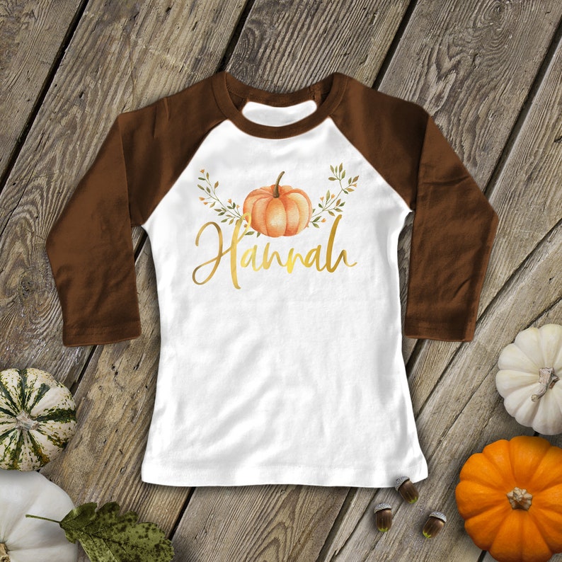 Pode incluir: Uma camiseta de beisebol branca e marrom com uma ab&oacute;bora dourada e o nome "Hannah" estampado na frente.
