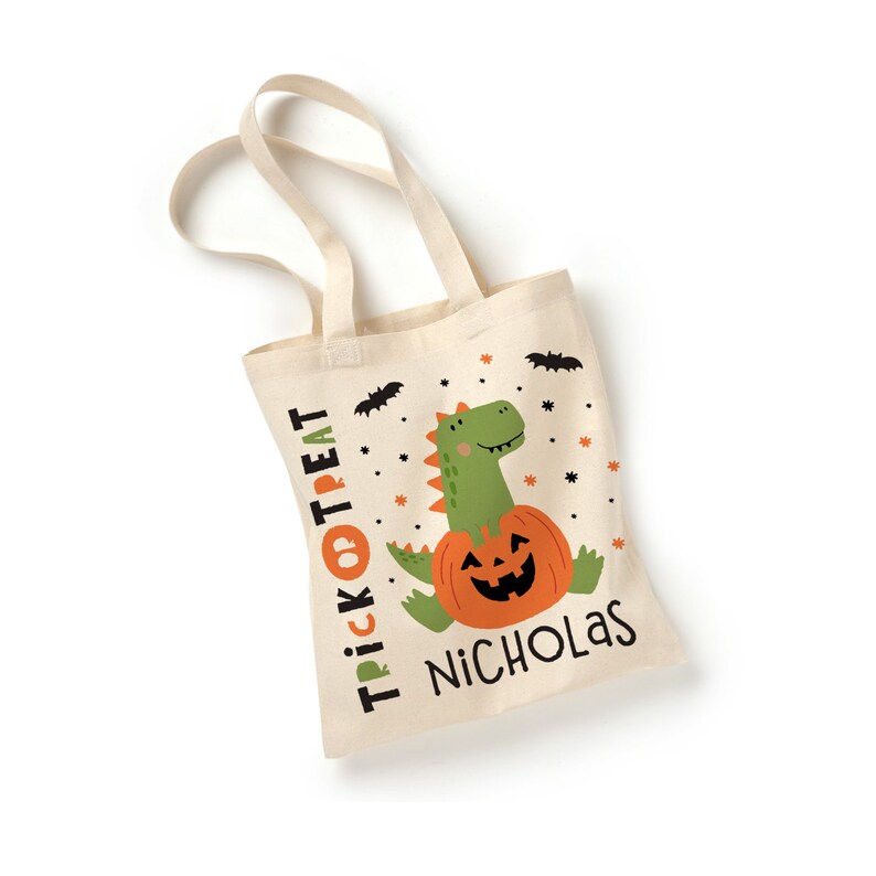 K&ouml;nnte beinhalten: Eine beige Stofftasche f&uuml;r S&uuml;&szlig;es oder Saures mit einem gr&uuml;nen Dinosaurier, der eine ausgeh&ouml;hlte K&uuml;rbislaterne h&auml;lt. Die Tasche hat den Text "Trick or Treat" und den Namen "Nicholas" aufgedruckt.