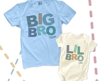 May include: Two t-shirts with the words "Big Bro" and "Lil Bro" in colourful patchwork letters. The larger shirt is light blue and says "Adam" below the text. The smaller shirt is cream and says "Cam" below the text.