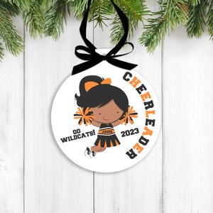 May include: White round ornament with a black and orange cheerleader design. The cheerleader is holding pom-poms and has a black ponytail. The text on the ornament reads "Cheerleader", "Go Wildcats!" and "2023".