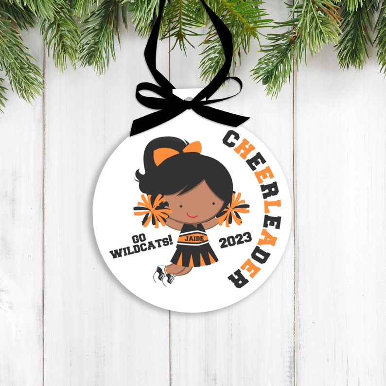 May include: White round ornament with a black and orange cheerleader design. The cheerleader is holding pom-poms and has a black ponytail. The text on the ornament reads "Cheerleader", "Go Wildcats!" and "2023".