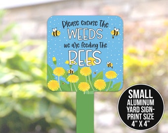May include: A blue and yellow metal garden sign with the text "Please excuse the WEEDS we are feeding the BEES". The sign features cartoon bees and dandelions. The sign is 10 cm by 10 cm.