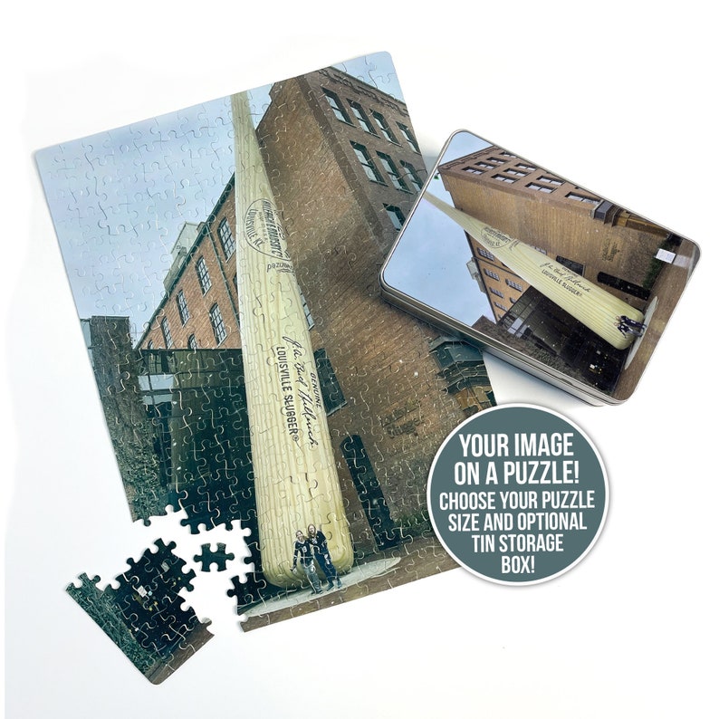 May include: A jigsaw puzzle featuring a large wooden baseball bat standing in front of a brick building. The bat is labeled "The Big Hiller" and "Louisville Slugger". The puzzle is in a metal tin box.