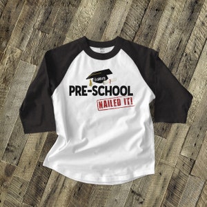 May include: White and black baseball tee shirt with a graduation cap and diploma graphic. The text "PRE-SCHOOL NAILED IT!" is printed on the shirt. The name "Garrett" is on the graduation cap.