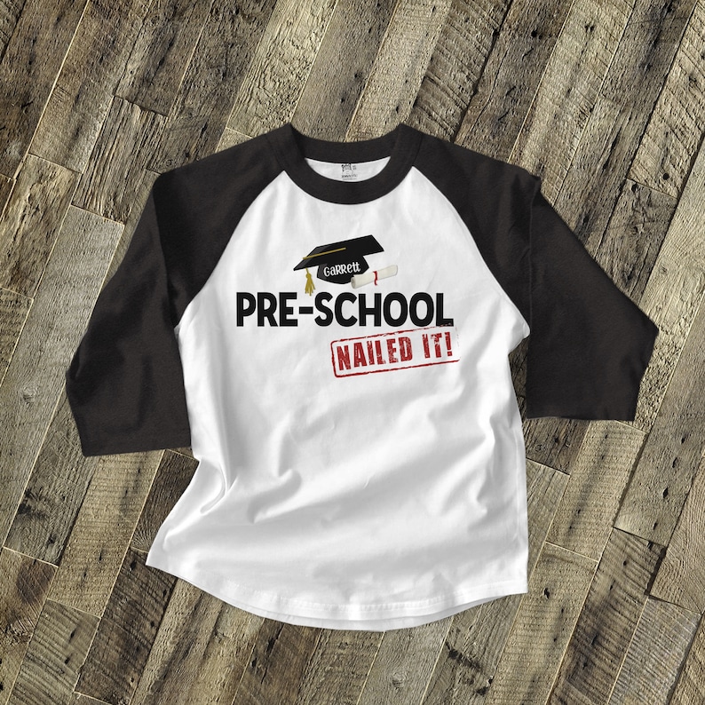 May include: White and black baseball tee shirt with a graduation cap and diploma graphic. The text "PRE-SCHOOL NAILED IT!" is printed on the shirt. The name "Garrett" is on the graduation cap.