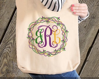 May include: A white canvas tote bag with a purple, green, and gold monogram surrounded by a wreath of Mardi Gras beads. The text "CHOOSE HEAVYWEIGHT OR VALUE BAG" is at the bottom of the image.