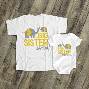 May include: Two white t-shirts featuring grey and yellow elephant designs. The larger shirt reads "Big Sister Jayda" and the smaller shirt reads "Baby Sister Ashiyana".