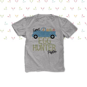 May include: A grey t-shirt with a blue truck design carrying Easter eggs. The text on the shirt says "Expert Egg Hunter Payton". The design includes a bunny driving the truck, ideal for Easter celebrations.