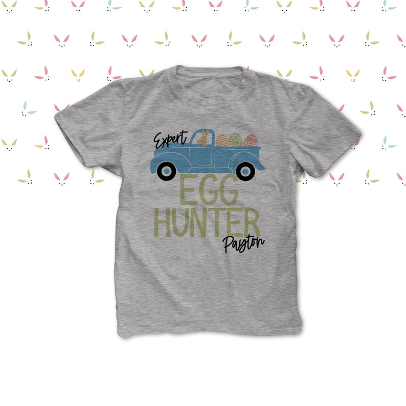 May include: A grey t-shirt with a blue truck design carrying Easter eggs. The text on the shirt says "Expert Egg Hunter Payton". The design includes a bunny driving the truck, ideal for Easter celebrations.