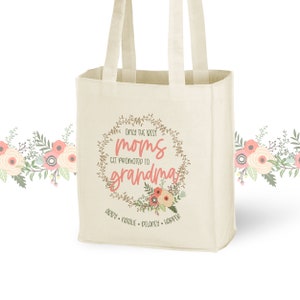 May include: A white canvas tote bag featuring a floral wreath design with the text "Only the best moms get promoted to grandma". The wreath is brown with pink flowers and green leaves. The text is in pink and brown. The names "Abby, Natalie, Delaney, Harper" are printed below the wreath.